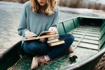 Portrait of young beautiful woman sitting on a boat and reading a book. She is bookworm and she choose between few books. Close up photo, no face shown.