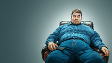 Portrait Of Fat Caucasian Man Wearing Jeanse And Whirt Sitting In A Brown Armchair Isolated On Gradient Grey Background. Emotional Watching TV And Changing Channels, Laughting. Overweight, Carefree.