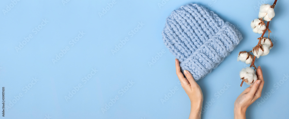 Flat lay fashionable blue knitted winter hat in female hands cotton flowers on blue background top view. Stylish woolen hat concept of winter accessories for the cold. Advertising shopping winter sale - obrazy, fototapety, plakaty 