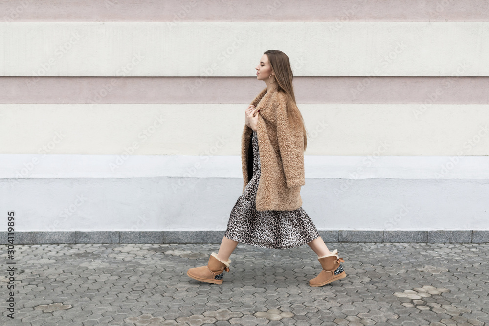 artificial fur coat, long leopard print dress, ugg boots, a small shoulder  bag, a beautiful white model girl with long hair poses cheerfully walks on  the street near the wall Stock Photo |
