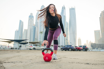 Beautiful  brunette fitness model wearing a black long sleeve top and purple about to grab a red kettlebell on the ground a with a cityscape background on a bright sunny day