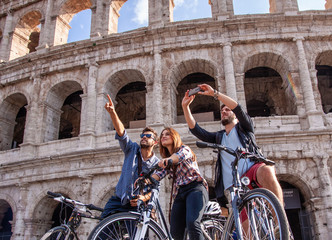 Wall Mural - Three happy young friends tourists with bikes at Colosseum in Rome taking pictures and selfies with smartphone