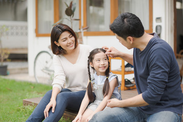 Young Asian family love, father mother and daughter sitting talking smiling at grass outdoor in front of home. girl looking at father which smiling and felling happy. background is white house.