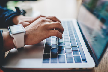 side view of female hands with smartwatch keyboarding text on modern laptop in search information fo