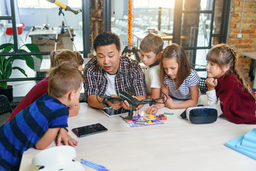 In physics and mechanics lesson young Asian teacher demonstrates quadcopter for caucasian pupils at class in modern smart school. Science, drone, engineering and future concept.