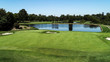 Golf course fairway, water hazard pond, sand bunkers, green with flag, lush green grass surrounded by trees against blue sky