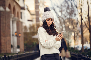 Wall Mural - Elegant girl walk in a winter city. Woman in a white knited sweater. Beautiful lady with dark hair. Girl use the phone