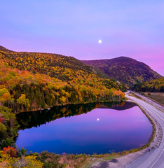 Aerial drone view of fall foliage in mountains with reflection of pink sky and moon in pond