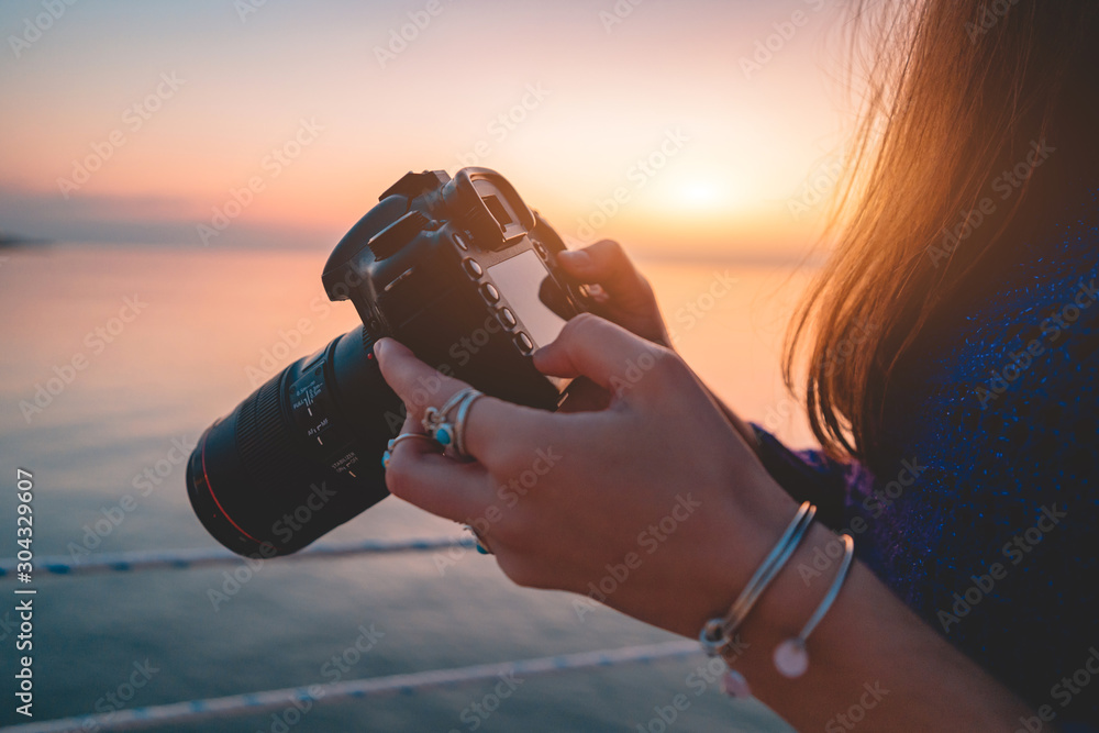 Woman photographer holds dslr camera during taking photos sea at sunset - obrazy, fototapety, plakaty 