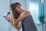 Happy attractive caucasian joyful brunette woman in bath towel dries hair with hair dryer in bathroom after shower at home.