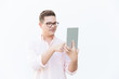 Focused nerdy guy in eyeglasses using tablet, looking at screen. Young man in glasses standing isolated over white background. Digital communication concept