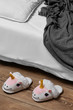 Subject shot of plush house slippers made in the form of white and pink smiling unicorn. The slippers are next to the bed with white linen, pillow and a gray plaid on it. 