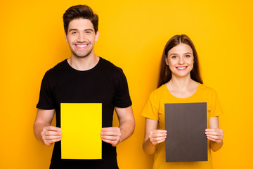 Sticker - Photo of cheerful cute nice couple of two spouses presenting you two pieces of paper held with hands with empty space on them isolated over vibrant shiny bright color background in black t-shirt