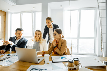 Group of a young office employees dressed casually in the suits having some office work at the large meeting table in the bright sunny room