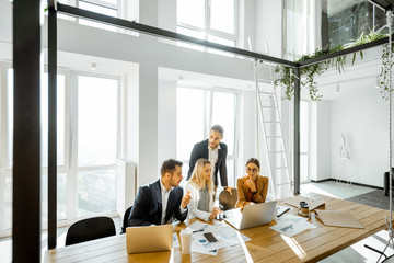 Group of a young office employees having some office work at the large meeting table, wide view on the spacious room with large windows