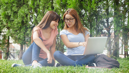 Wall Mural - Asian teenager girl lying on the grass with her caucasian friend using laptop. Happiness and lightheartedness concept