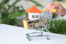 The Hand Is Pushing The Cart.Orange Roof House And Coins In A Wheelchair. House Trading Ideas Saving Money To Buy A Family Home Real Estate Investment, Mortgage. Shopping Requires A Lot Of Money.