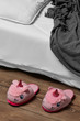 Subject shot of plush house slippers made in the form of pink smiling unicorn. The slippers are next to the bed with white linen, pillow and a gray plaid on it. 