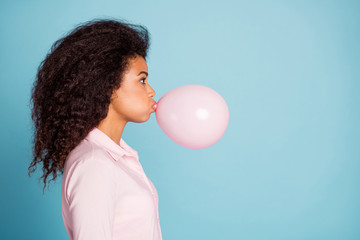 Wall Mural - Closeup profile photo of pretty dark skin lady holding big air balloon in mouth making decorations for surprise party wear pink shirt isolated blue color background