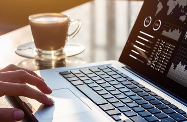 Person working on laptop computer with business analytics and financial report data with metrics and charts on screen at modern office workplace with sunset light and coffee