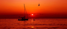 Silhouette Of A Sailboat Between The Sunrise Under A Red Sky With Flying Seagulls.