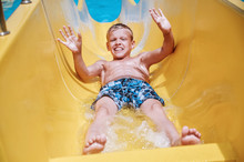 Sporty Boy Waves His Arms And Starts From A Yellow Slide Down To The Pool In Sunny Good Weather On Vacation