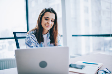 Wall Mural - positive female employee in casual wear sitting at desktop reading information on web page via lapto