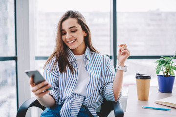 Wall Mural - cheerful young woman checking new feed and content in social networks via smartphone during coffee b