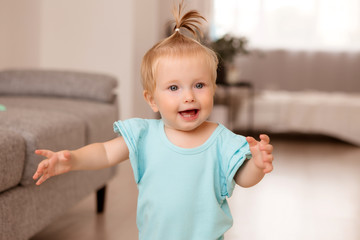 Wall Mural - healthy baby girl in a blue bodysuit in a room next to a gray sofa is learning to walk. the comfort of the home. the child smiles