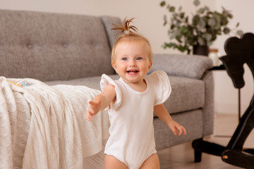 Wall Mural - healthy baby girl in a white bodysuit in a room next to a gray sofa is learning to walk. the comfort of the home. the child smiles