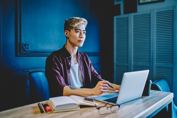 Thoughtful asian hipster student thinking on creative ideas for development own startup project sitting at table desktop with laptop computer for productive work, concept of distance job indoors