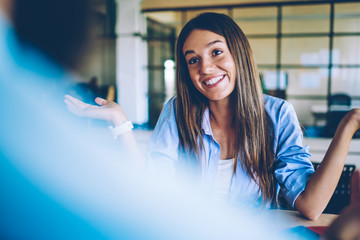 Wall Mural - Positive smiling woman 20s with brunette hair sitting at table and talking with male friend in front, back view of successful caucasian hipster girl spending time with man and discussing ideas