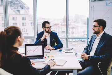 Working team of business people sitting at table in meeting room with modern computers, clever workers analyzing and discussing new strategy development of company using graphs and feeling successful