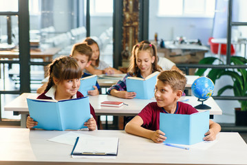 Handsome boy and pretty girl of school sitting together at the desk, look each other and smile while reading book on lesson. Education and pupils concept.