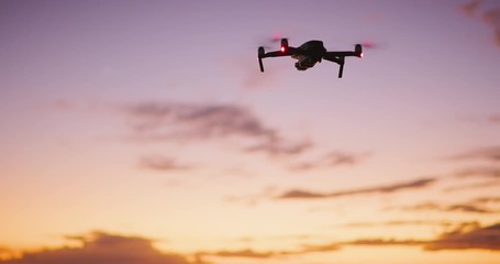 orbiting view of a quadcopter drone silhouette hovering and flying at sunset with a purple yellow sk