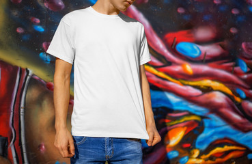 Wall Mural - Presentation of a white T-shirt on a young guy, against the background of graffiti, front view.
