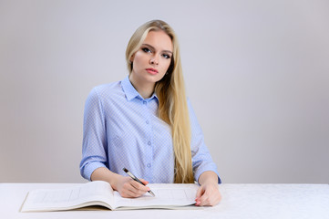 Concept cute model student secretary works sitting at a table. Close-up portrait of a beautiful blonde girl with excellent makeup with long smooth hair on a white background in a blue shirt.