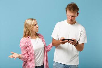 Wall Mural - Young couple two friends guy girl in white pink empty t-shirts posing isolated on pastel blue background. People lifestyle concept. Mock up copy space. Playing video game with mobile phones, swearing.