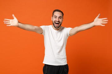 Wall Mural - Joyful pleasant young man in casual white t-shirt posing isolated on orange wall background. People sincere emotions lifestyle concept. Mock up copy space. Standing with spreading outstretched hands.