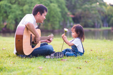 Wall Mural - Asian father is playing guitar nearly with his daughter sitting and handling white flower in the background of green park with happiness moment, concept of activity in family lifestyle.