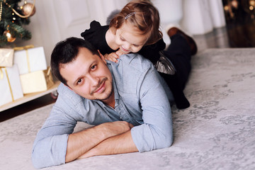 A handsome man plays with his funny daughter near the Christmas tree at home. Dad and her smiling baby