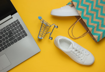 Wall Mural - Online Shopping concept. Fashion studio shot. Laptop, white sneakers in paper bag,  shopping trolley on yellow background. Top view