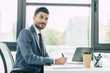 © LIGHTFIELD STUDIOS - smiling businessman writing in notebook and looking away while sitting at workplace