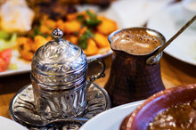 Turkish Coffee And Traditional Embossed Metal Cup Placed On Table. Food Blurred At Foreground And Background.