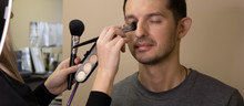 Young Professional Makeup Artist Girl Holding Eyeshadows Kit And Cosmetic Brushes And Applying Makeup On An Attractive Male Face.