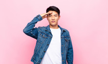 Young Chinese Man Greeting The Camera With A Military Salute In An Act Of Honor And Patriotism, Showing Respect Against Flat Color Wall