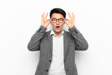 Young Chinese Man Feeling Shocked, Amazed And Surprised, Holding Glasses With Astonished, Disbelieving Look Against Flat Color Wall