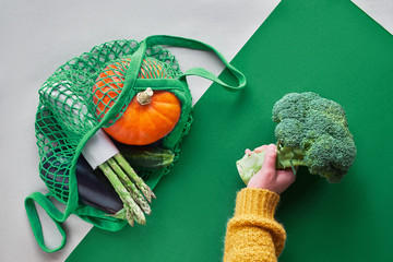 Eco friendly zero waste flat lay with hands holding broccoli and string bag with orange pumpkin and green asparagus packed in craft paper. Top view on two color brown and green paper background.