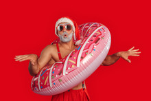 Christmas Freestyle. Young Santa Claus Bare Muscular Upper Body In Hat And Sunglasses Standing Isolated On Red In Swim Ring Posing To Camera Playful