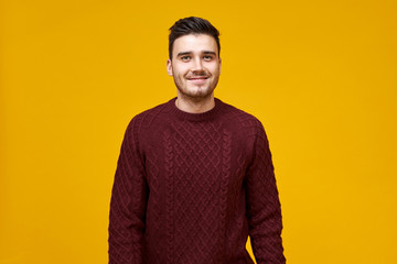 Wall Mural - Studio shot of handsome cheerful young man with stylish haircut and dimpled smile posing isolated against blank yellow studio wall background, dressed in cozy maroon sweater, having confident look